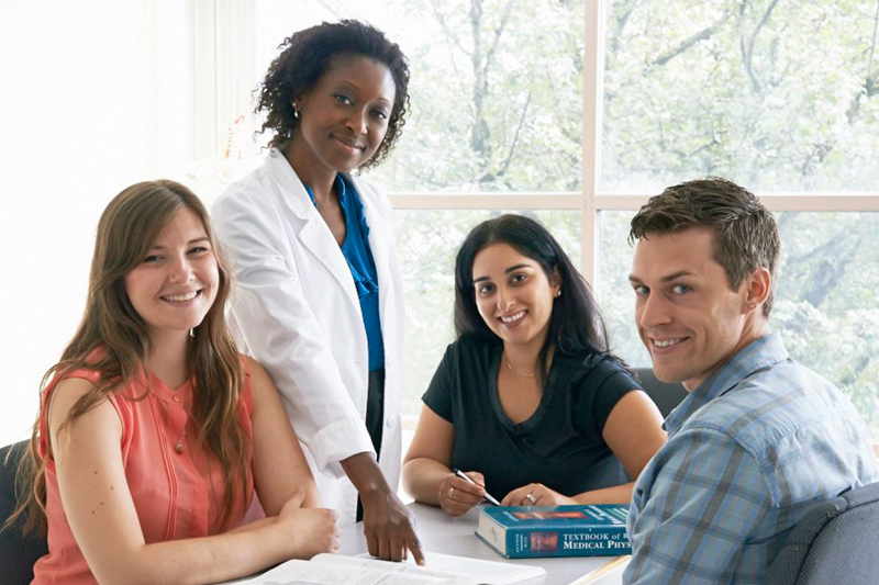 four students - three female and one male student - smiling and pointing to a textbook